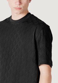 Maglietta nera a maniche corte con un motivo a diamante testurizzato, scollo rotondo e una finitura del tessuto liscia. Cuciture visibili sulle maniche.