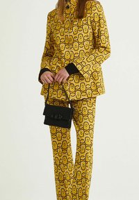 Costume jaune à motifs géométriques, avec des accents noirs sur les poignets. Un petit sac noir à texture est tenu à la main.