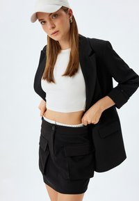 Zwarte blazer over een witte cropped tank, gecombineerd met een zwarte mini-jurk met twee voorzakken en een gerimpelde tailleband. Witte accenten.