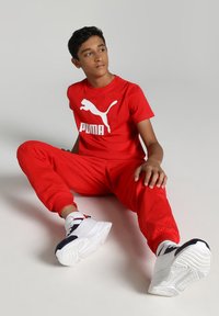 Červené tričko s krátkym rukávom značky Puma s bielym logom, v kombinácii s červenými teplákmi. Biele tenisky s čiernymi akcentmi a gumovou podrážkou.