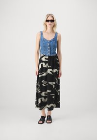 Top court en denim avec des boutons associé à une jupe maxi noire présentant un motif abstrait beige. Le modèle porte des chaussures noires à enfiler.