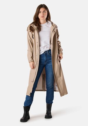 Beige raincoat with a hood, long length, adjustable cuffs, front buttons and pockets. Worn over a white t-shirt and blue jeans with black boots.