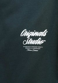 Tissu vert foncé avec une surface texturée, présentant un texte brodé blanc : "Originals Studio™" et des détails de marque supplémentaires en dessous.