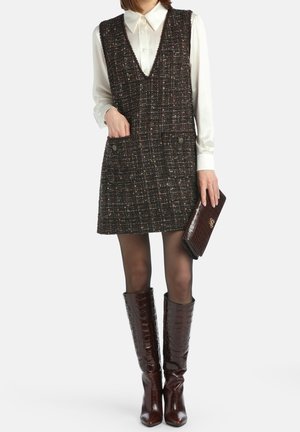 Frau trägt ein ärmelloses Tweed-Kleid über einem weißen Hemd mit Kragen, hält eine dunkelbraune Clutch und trägt kniehohe braune Stiefel.
