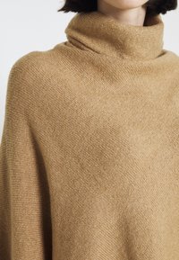 Femme portant un pull à col roulé en maille couleur camel, montrant la texture et les détails des plis près du cou et des épaules.