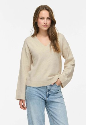 Femme aux longs cheveux bruns portant un pull beige à col en V et un jean bleu clair, debout avec une main dans la poche sur un fond blanc.