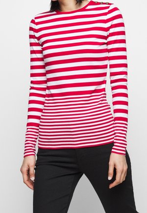 Osoba ubrana w czerwono-białą bluzkę w poziome paski z długimi rękawami, złotymi guzikami na ramieniu oraz czarne spodnie.