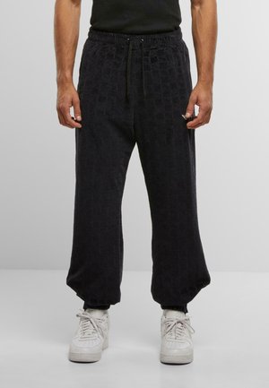 Uomo che indossa pantaloni da jogging neri con texture, polsini elasticizzati, vita con cordino e sneakers bianche, in piedi davanti a uno sfondo grigio uniforme.