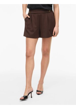 Femme portant un short taille haute marron foncé avec des plis et des mules à talons hauts noires à bout ouvert, debout, une main dans la poche.