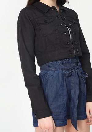 Femme portant une veste noire courte à boutons par-dessus un haut rayé et un short taille haute bleu style paperbag noué avec une ceinture en tissu.