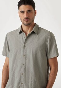 Camisa de manga corta en tejido verde claro con un patrón texturizado, cierre de botones en la parte delantera y un cuello clásico.