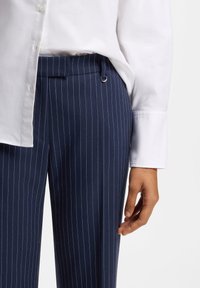 Pantalon bleu marine à fines rayures, confectionné dans un tissu lisse, avec une boucle latérale et une taille ajustée, accompagné d'une chemise blanche à boutons.