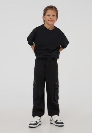 T-shirt nero a maniche corte abbinato a pantaloni cargo neri larghi con tasche con zip, indossato con sneakers bianco e nere.