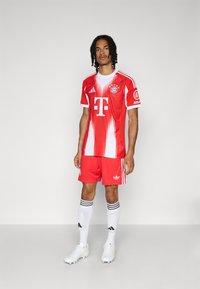 Maillot de football rouge avec des rayures blanches portant un logo, associé à un short rouge assorti, des chaussettes blanches avec des rayures noires et des chaussures blanches.