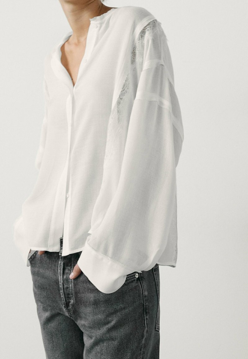 Personne portant un chemisier blanc ample boutonné avec de longues manches bouffantes et un jean gris foncé, debout les mains dans les poches.