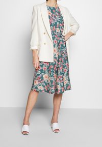 Abito verde floreale con motivi rosa e bianchi, abbinato a un blazer bianco oversize e scarpe slide bianche. L'abito ha la vita cinte.