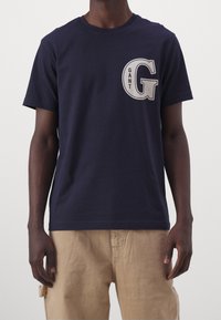Camiseta de algodón de color azul marino con cuello redondo, que presenta una gran letra blanca "G" y el texto "GANT" superpuesto en el lado izquierdo del pecho. Mangas cortas.