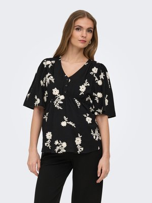 Frau mit langen Haaren, die eine schwarze V-Ausschnitt-Bluse mit weißer Blumenstickerei und weiten Ärmeln trägt, kombiniert mit schwarzen Hosen, steht vor einem grauen Hintergrund.