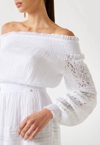Abito bianco scollato sulle spalle con texture a pieghe, busto arricciato e maniche a sbuffo con pizzo. Accenti decorativi in vita.
