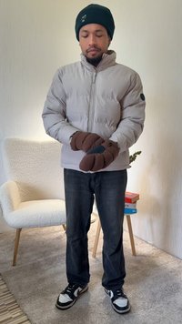 Casaco acolchoado cinza claro com gola alta, luvas castanhas, jeans escuros e sapatilhas pretas e brancas, sobre um fundo neutro.