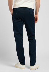 Pantalons chino bleu marine à coupe slim, avec deux poches arrière et une texture lisse, associés à des chaussures de couleur claire.