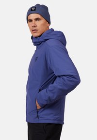 Lila Kapuzenjacke mit Reißverschlusstaschen und Logo auf der Brust, kombiniert mit einer blauen Strickmütze, strukturiertem Stoff und lockerer Passform.