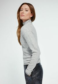 Kobieta z długimi brązowymi włosami, ubrana w szary sweter z golfem i czarne skórzane spodnie, stojąca z rękami w kieszeniach i patrząca w obiektyw.