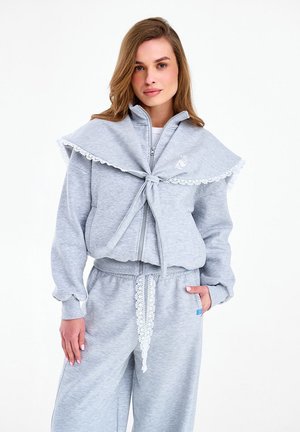 Kobieta ubrana w jasnoszarą bluzę na zamek z detalem zawiązywanego kołnierzyka oraz dopasowane spodnie dresowe z koronkowym troczkiem, stojąca z jedną ręką w kieszeni.