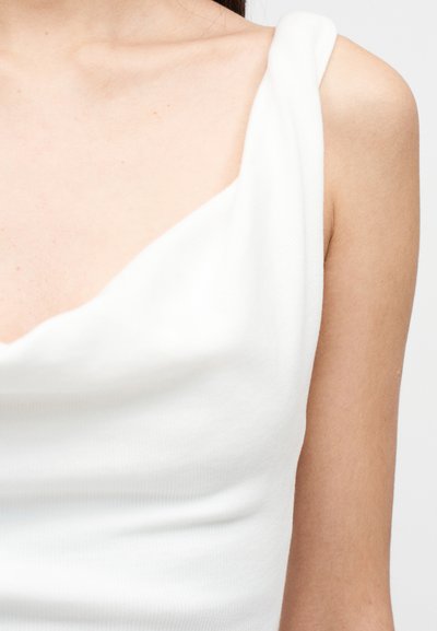 Λευκό αμάνικο τοπ με στρογγυλεμένο λαιμόκομμα και υφασμάτινη υφή. Διαθέτει απαλές πτυχώσεις γύρω από τους ώμους και εφαρμοστή σχεδίαση.