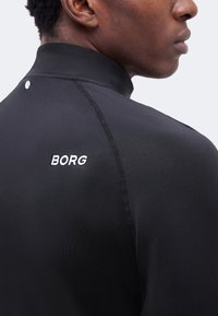 Νέος άνδρας που φοράει μαύρο αθλητικό τοπ με ψηλό γιακά, με ορατές ραφές και την επιγραφή "BORG" σε λευκό στο πάνω μέρος της πλάτης.