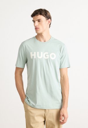 Ung mand med kort brunt hår iført en lys grøn HUGO T-shirt og beige bukser, stående med den ene hånd i lommen, og ser til venstre.