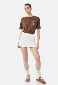 Camiseta marrón con estampado gráfico, minifalda blanca con botones laterales, calcetas crema hasta la rodilla y zapatillas color tostado con cordones.