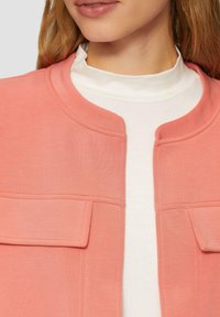Helle rosa Jacke mit runder Ausschnitt, ausgestattet mit zwei Fronttaschen, glatter Textur und leichtem Glanz über einem weißen Rollkragenpullover.