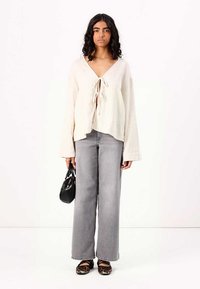 Light beige, long-sleeve blouse with a tie-front neckline, paired with high-waisted, wide-leg grey jeans and black shoes, holding a black handbag.