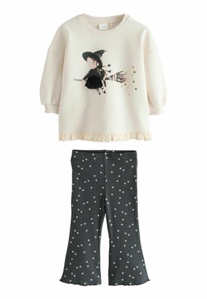 Gräddfärgad sweatshirt med en häxa-illustration och volangkant, tillsammans med mörkgrå utsvängda byxor med vita stjärnmönster.