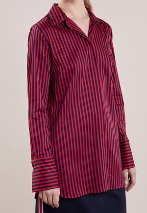 Femme portant une blouse à rayures verticales rouges et navy à manches longues, avec de larges poignets et un col, assortie d'une jupe sombre.