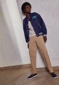 Woman with short curly hair wearing navy jacket, beige pants, and navy slip-on shoes, standing against a textured white wall indoors.