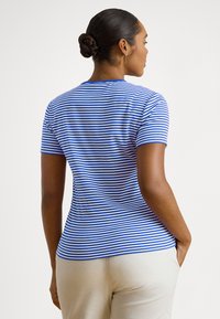 Γυναίκα με σκούρα μαλλιά μαζεμένα σε κότσο, φορώντας μπλε και άσπρη ριγέ κοντομάνικη μπλούζα και ανοιχτόχρωμο παντελόνι, που στέκεται με την πλάτη προς τη κάμερα μπροστά σε απλό φόντο.