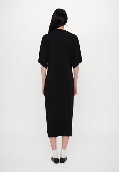 Robe noire midi avec des manches larges, design simple et une fente à l'arrière, en tissu doux. Le modèle se tient debout avec des chaussettes et des chaussures claires.