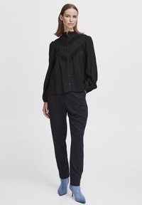 Svart blus med knappar och puffärmar, med texturerade paneler och en subtil lyster, parat med mörka byxor och blå ankelstövlar.