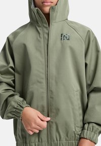 Oliivivihreä hupullinen takki, jossa on elastiset hihansuut ja kankaan vyötärö. Varustettu kaksisuuntaisella vetoketjulla ja pienellä logolla rinnassa. Silkkinen kangas.