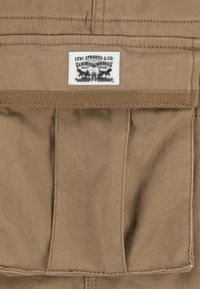 Tela caqui de textura suave con dos bolsillos frontales. Una etiqueta blanca de Levi Strauss & Co. con texto negro es visible en la parte superior.