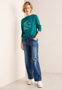 Teal lange mouwen sweatshirt met grafische print, gecombineerd met blauwe wijde jeans en witte sneakers, staande tegen een neutrale achtergrond.