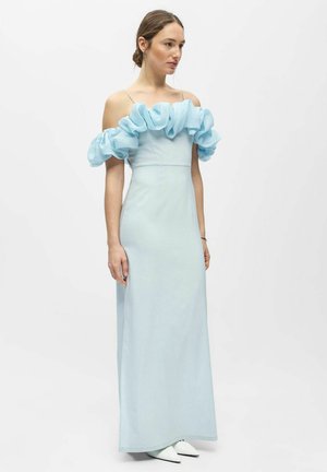 Vrouw staand, gekleed in een lichtblauwe jurk tot op de vloer met off-shoulder gerimpelde mouwen en dunne schouderbandjes.