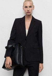 Černý pruhovaný blazer s ostrými klopami, se dvěma knoflíky a přední kapsou, doplněný velkou černou koženou taškou.
