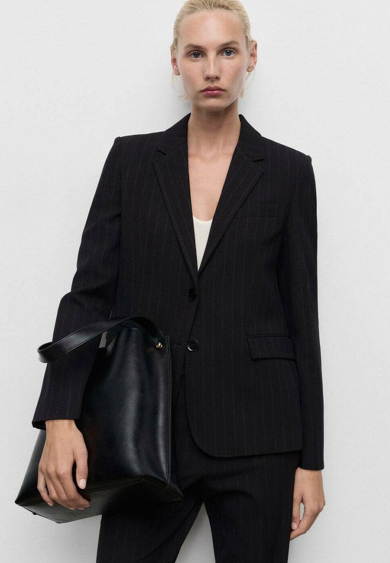 Černý pruhovaný blazer s ostrými klopami, se dvěma knoflíky a přední kapsou, doplněný velkou černou koženou taškou.