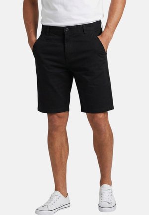 Homme portant un short noir arrivant aux genoux et des baskets blanches basses, debout les mains dans les poches sur un fond uni.