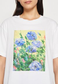 Bílý bavlněný tričko s barevným květinovým potiskem, na kterém jsou modré a růžové květiny na světlém žlutém pozadí. Krátké rukávy a kulatý výstřih.