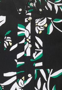 Chemise noire avec un motif floral comprenant des accents blancs, verts et jaunes. Col à boutons avec des boutons blancs sur le devant.