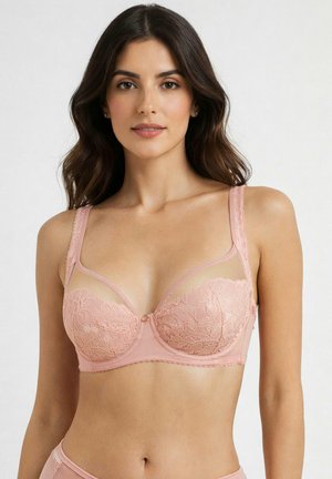 Kobieta z długimi ciemnymi włosami, ubrana w różowy koronkowy biustonosz z fiszbinami i przezroczystymi górnymi panelami, stojąca na tle jednolitego jasnego tła.